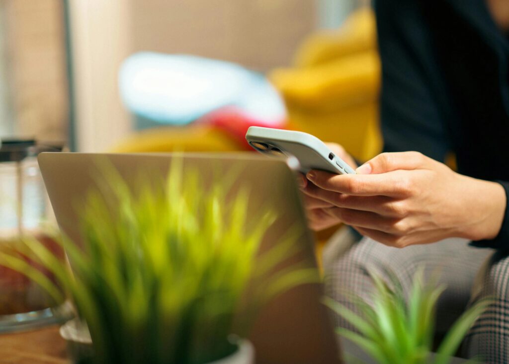 Eine Frau erledigt Multitasking auf ihrem Smartphone und Laptop, umgeben von grünen Pflanzen in einem gemütlichen Wohnzimmer.